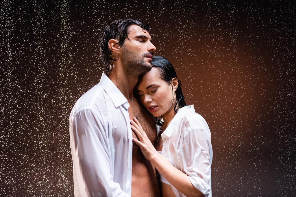 junges Paar in weißen Hemden steht unter Regen mit geschlossenen Augen vor dunklem Hintergrund - Foto, Bild