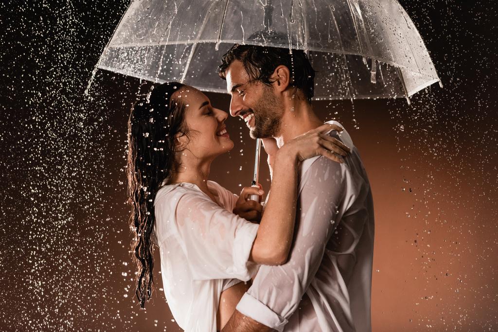 πλαϊνή άποψη του ευτυχισμένου ζευγαριού αγκαλιάζει ενώ στέκεται με ομπρέλα κάτω από τη βροχή σε σκούρο φόντο - Φωτογραφία, εικόνα