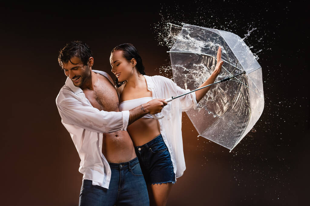 fröhliches Paar in nassen Hemden, mit durchsichtigem Regenschirm in der Nähe von Wasserspritzern auf dunklem Hintergrund - Foto, Bild