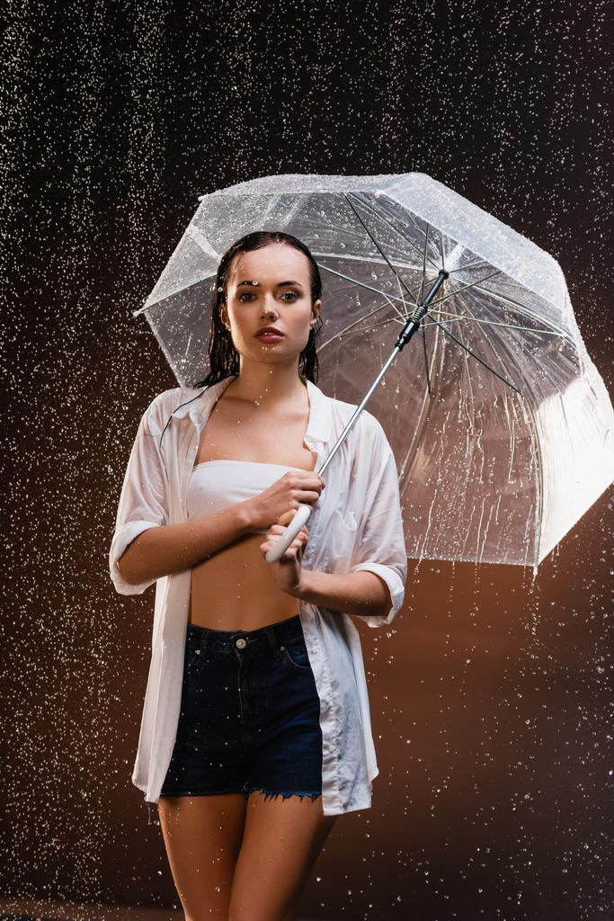 sinnliche, nasse Frau, die mit Regenschirm vor dunklem Hintergrund in die Kamera blickt - Foto, Bild