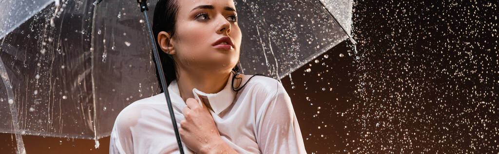 мокра жінка, дивлячись далеко, стоячи з дощем під прозорою парасолькою на темному фоні, банер
 - Фото, зображення