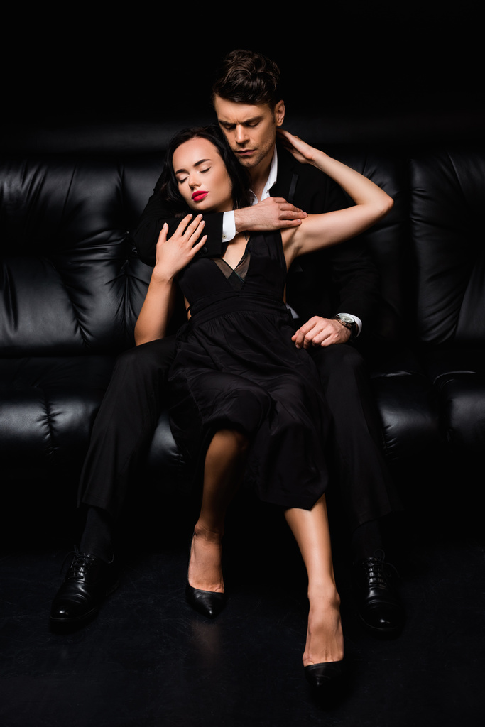 hombre con los ojos cerrados abrazando a mujer joven en vestido mientras está sentado en negro  - Foto, Imagen