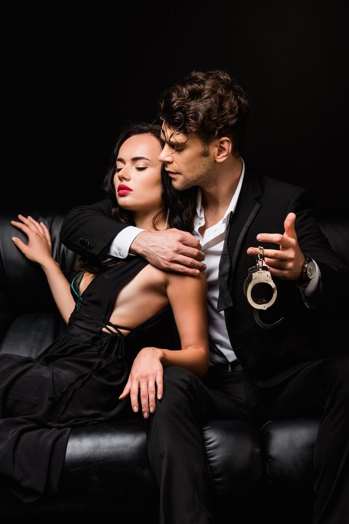 Mann hält Handschellen in der Nähe von sexy Frau im Kleid, während er auf Sofa isoliert auf schwarz sitzt - Foto, Bild