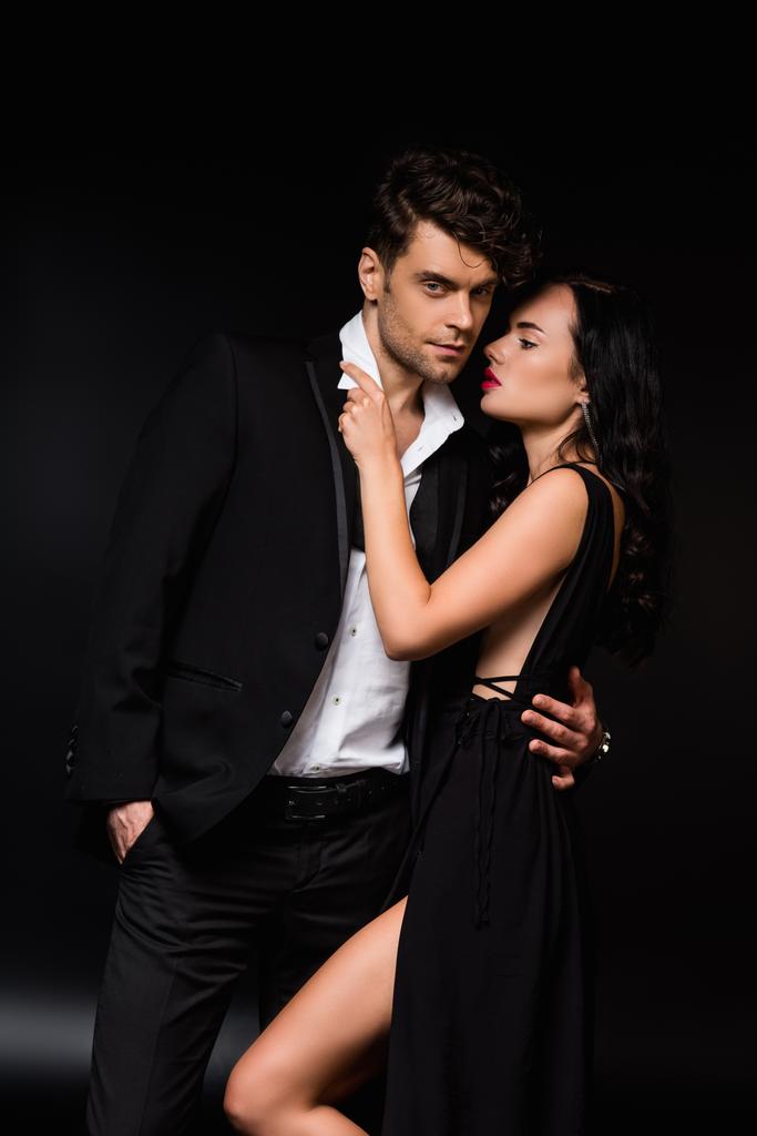 man standing with hand in pocket while hugging sensual woman in dress on black  - Photo, Image