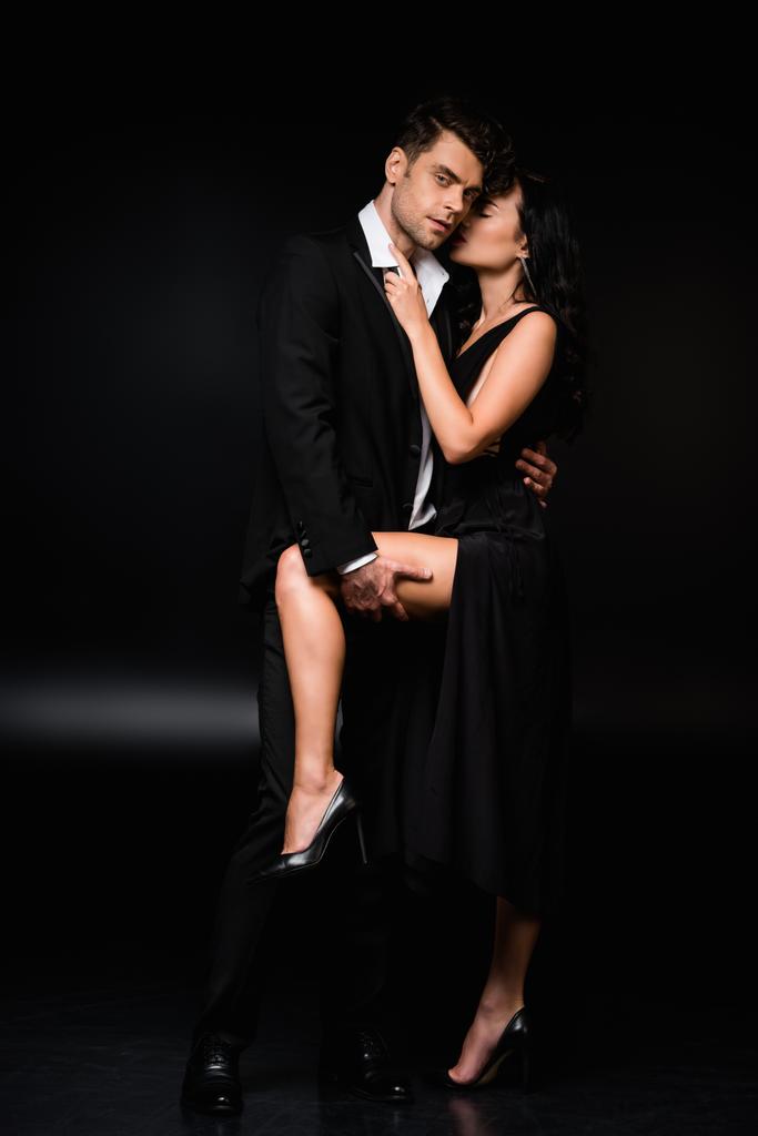 passionate man lifting leg of sexy woman in dress on black - Photo, Image