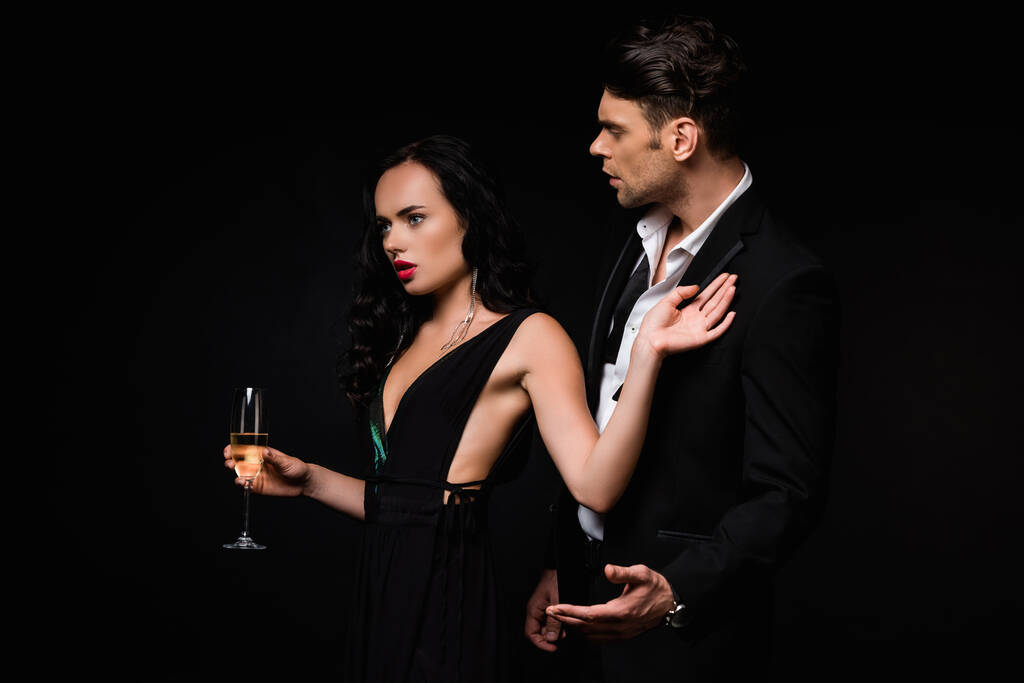 brunette woman in dress holding glass of champagne and pulling away man in suit isolated on black  - Photo, Image