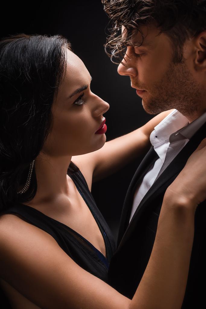 sexy mujer con labios rojos abrazando con hombre en traje aislado en negro  - Foto, imagen