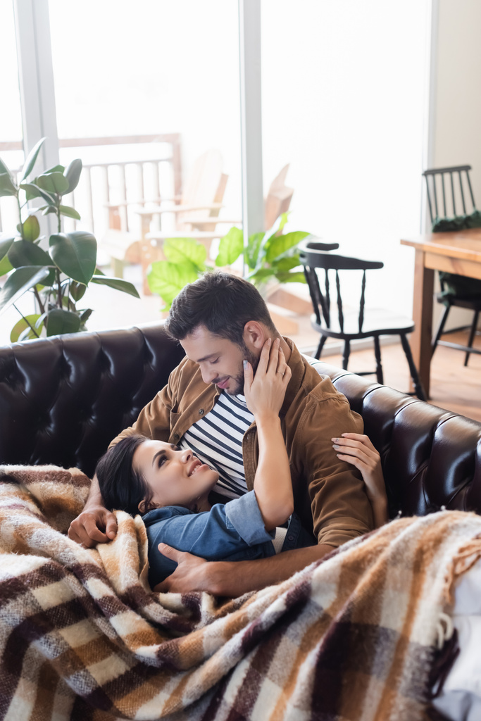 счастливая молодая пара обнимается, отдыхая на кожаном диване под уютным клетчатым одеялом - Фото, изображение