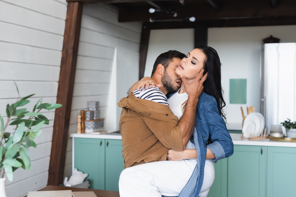 jonge man knuffelen en kussen nek van verleidelijke brunette vriendin in keuken - Foto, afbeelding
