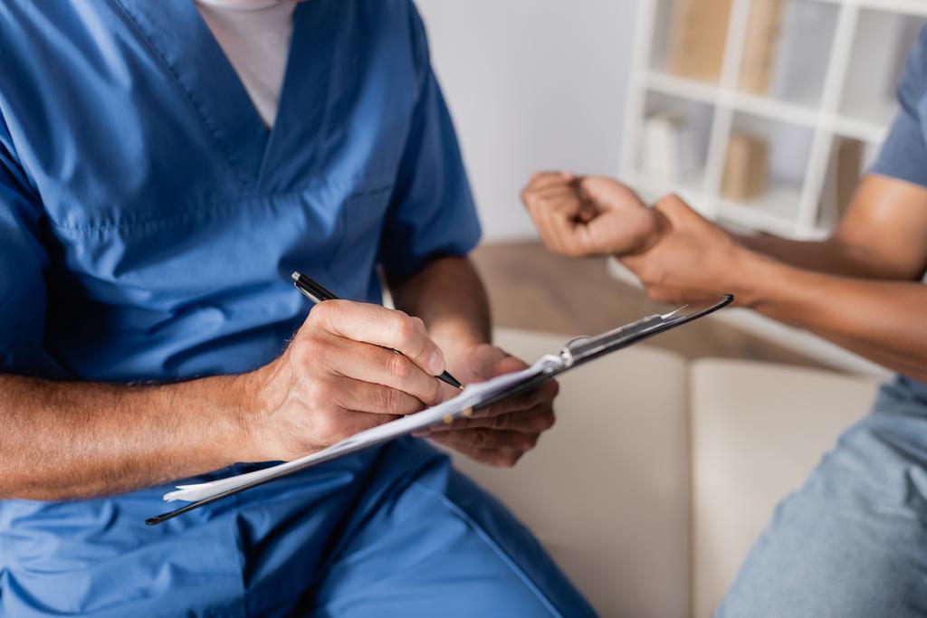 частичный взгляд врача, выписывающего рецепт рядом с раненым африканским американцем на размытом фоне  - Фото, изображение