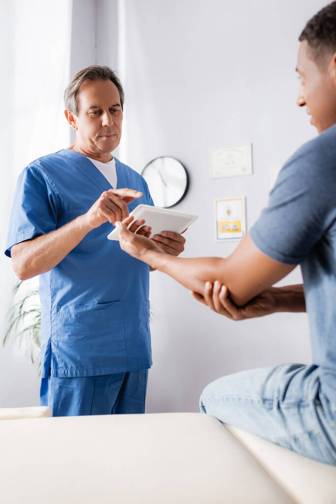зрелый врач держит цифровой планшет и указывает пальцем на африканского американского пациента на размытом переднем плане  - Фото, изображение