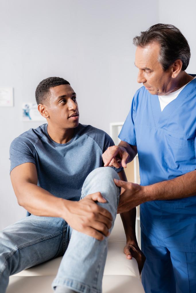 Dojrzały terapeuta wskazujący palcem na ranne kolano afrykańskiego mężczyzny w klinice - Zdjęcie, obraz