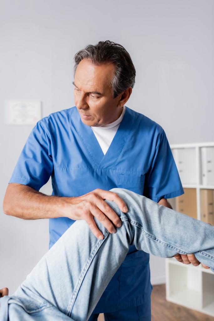masajista maduro trabajando con rodilla lesionada de paciente en clínica - Foto, Imagen