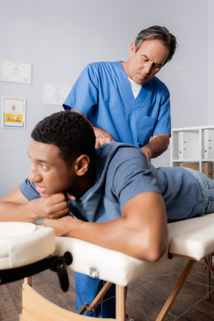 chiropratico di mezza età che lavora con un giovane paziente afroamericano in primo piano sfocato - Foto, immagini