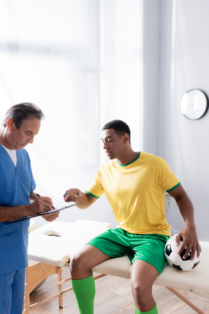 Arzt schreibt Rezept in der Nähe verletzter afrikanischer Fußballspieler sitzt mit Ball auf Massagetisch - Foto, Bild