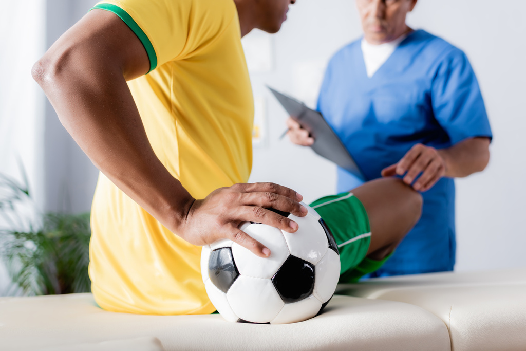 負傷したアフリカ系アメリカ人サッカー選手の切り裂かれた景色ぼやけた背景にクリップボードで医師の近くのマッサージテーブルの上にボールで座って - 写真・画像