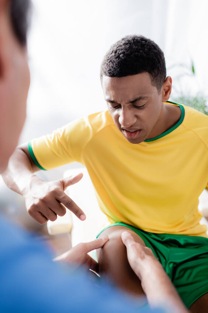 τραυματισμένος Αφροαμερικανός αθλητής που δείχνει με το δάχτυλο στο γόνατο κοντά στο γιατρό σε θολή πρώτο πλάνο  - Φωτογραφία, εικόνα