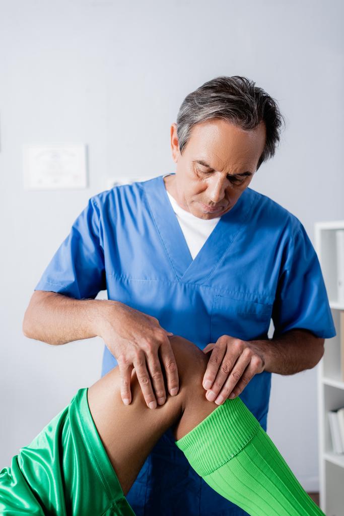 massagista de meia idade massageando joelho de esportista afro-americano na clínica  - Foto, Imagem