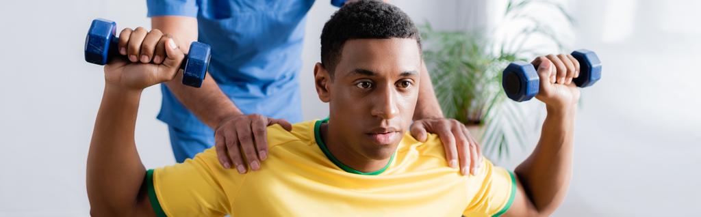 Африканский американский спортсмен с гантелями рядом с физиотерапевтом на размытом фоне, баннер - Фото, изображение