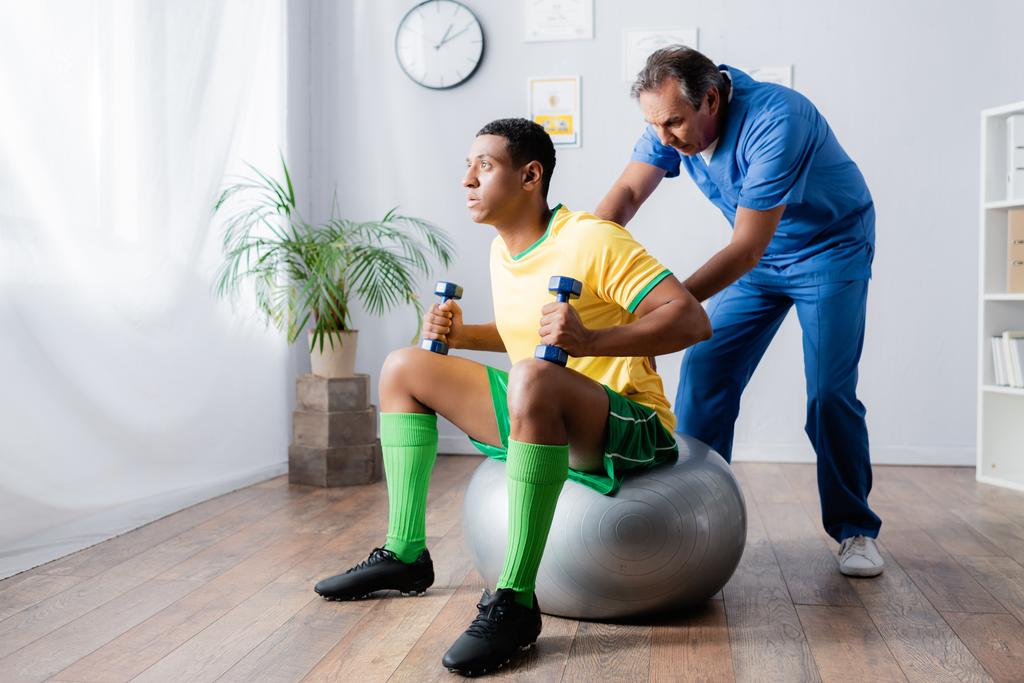un sportif afro-américain s'entraîne avec des haltères sur un ballon de fitness près d'un physiothérapeute - Photo, image