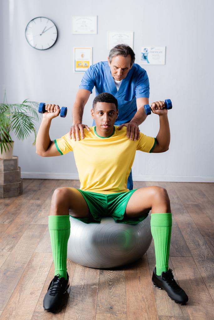 entraînement sportif afro-américain avec haltères sur balle de fitness près du physiothérapeute pendant la réadaptation  - Photo, image