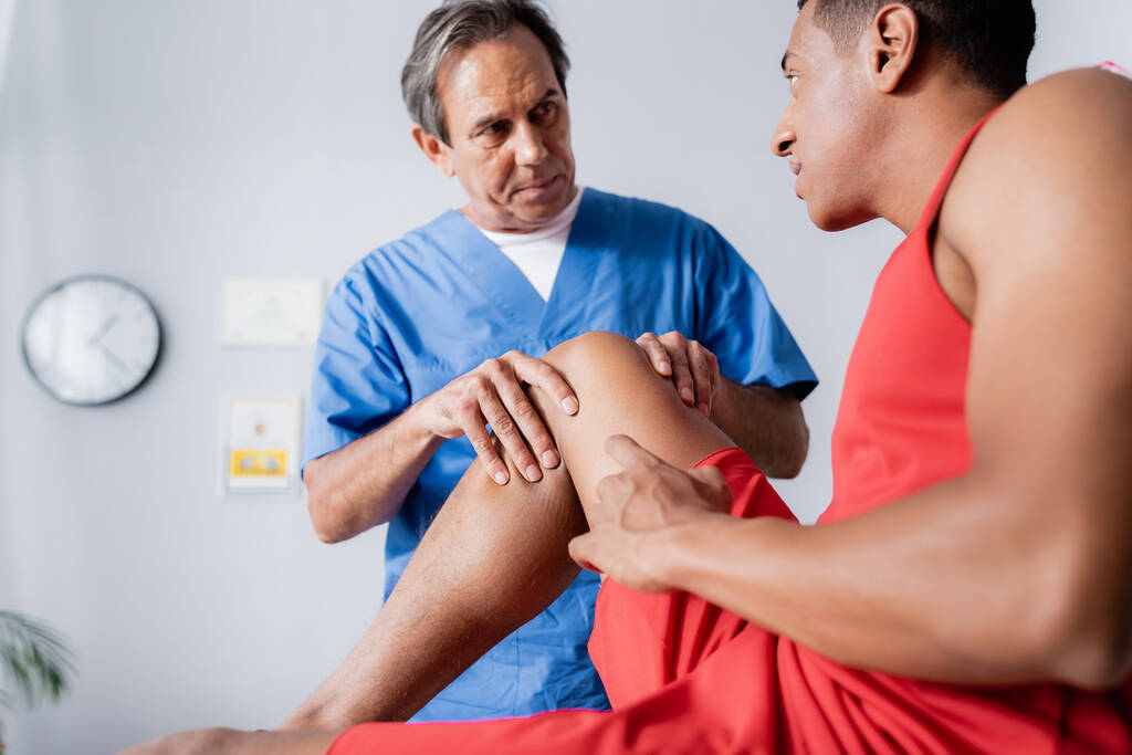 volwassen chiropractor werken met gewonde knie van Afrikaanse Amerikaanse man in sportkleding op wazig voorgrond  - Foto, afbeelding