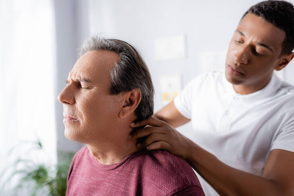 アフリカ系アメリカ人理学療法士がクリニックで成人男性の首をマッサージ - 写真・画像