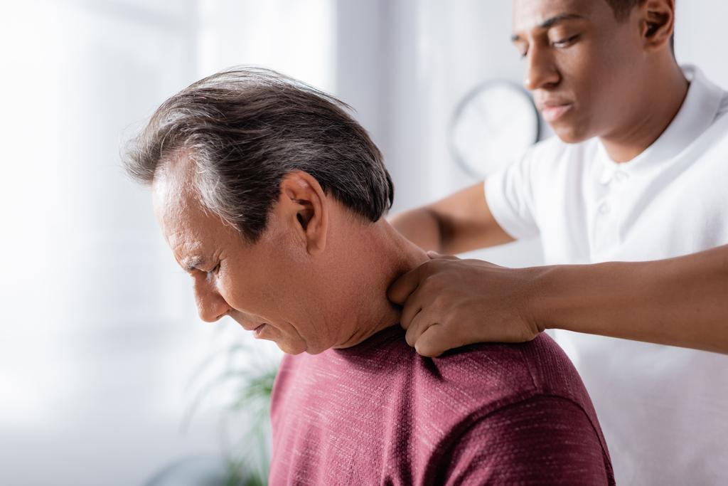 fisioterapista afroamericano che massaggia l'uomo di mezza età in clinica - Foto, immagini