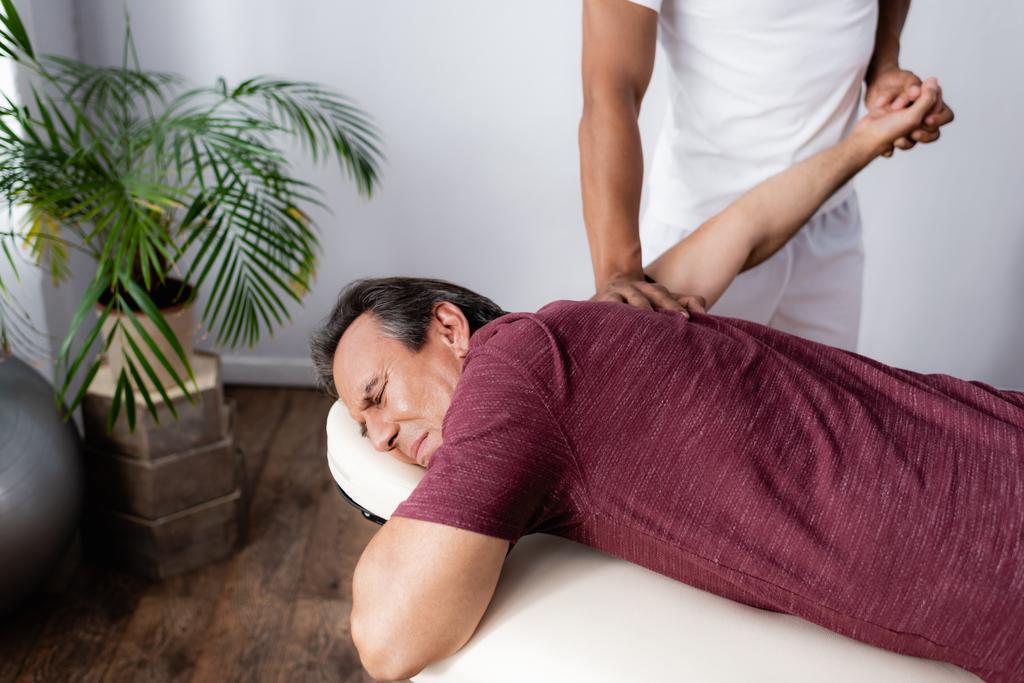 афроамериканський фізіотерапевт виправляє руку чоловіка середнього віку на масажному столі
 - Фото, зображення
