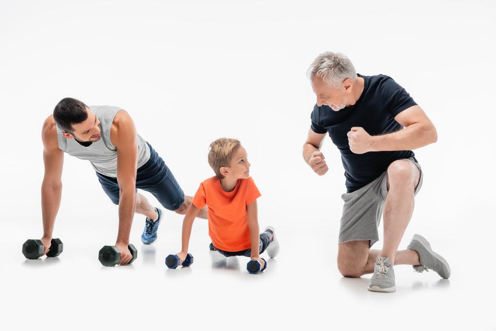 gelukkig grootvader demonstreren kracht in de buurt vader en kind doen push-ups met halters op wit - Foto, afbeelding