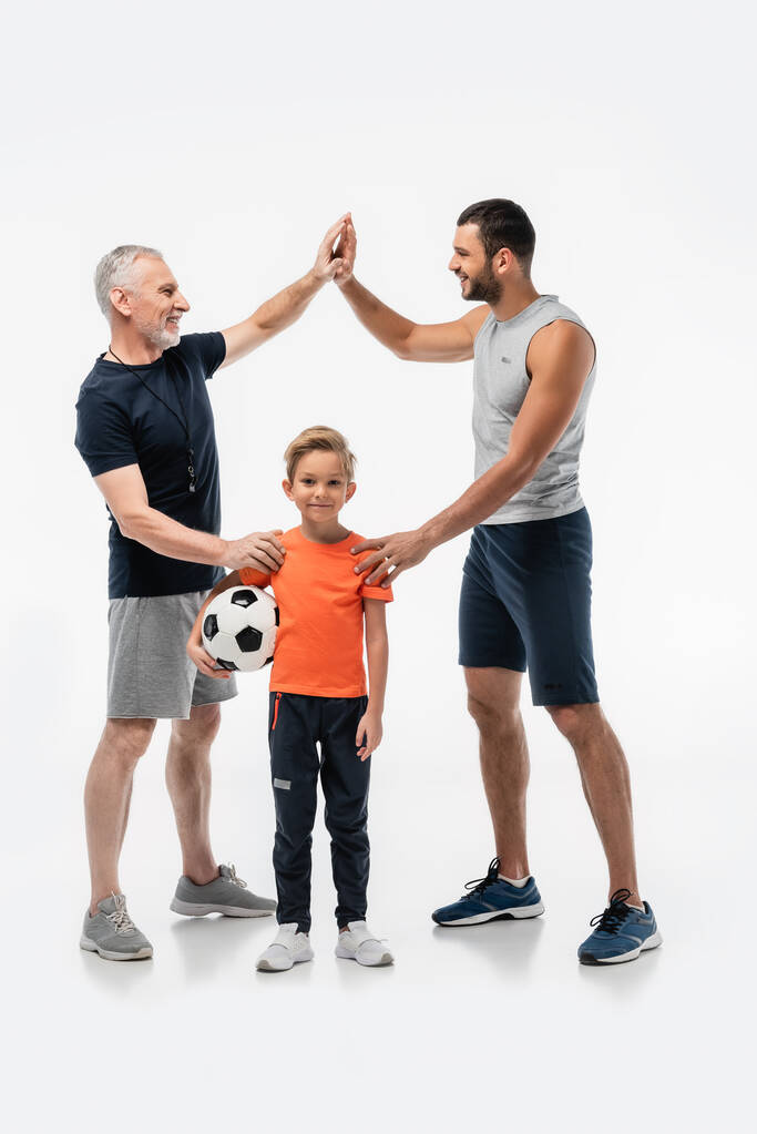 χαρούμενο αγόρι κρατώντας μπάλα ποδοσφαίρου και χαμογελώντας στην κάμερα κοντά στον μπαμπά και τον παππού δίνοντας κόλλα πέντε στα λευκά - Φωτογραφία, εικόνα