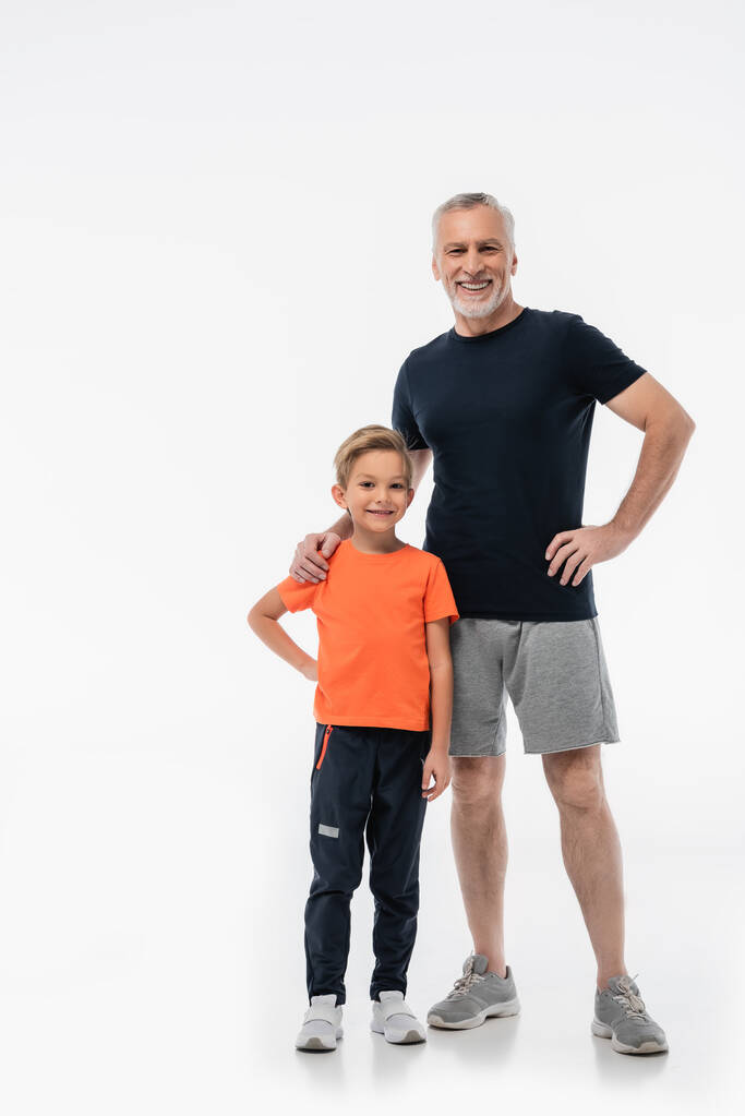 usmívající se dědeček a šťastný vnuk ve sportovním oblečení při pohledu na kameru na bílém - Fotografie, Obrázek