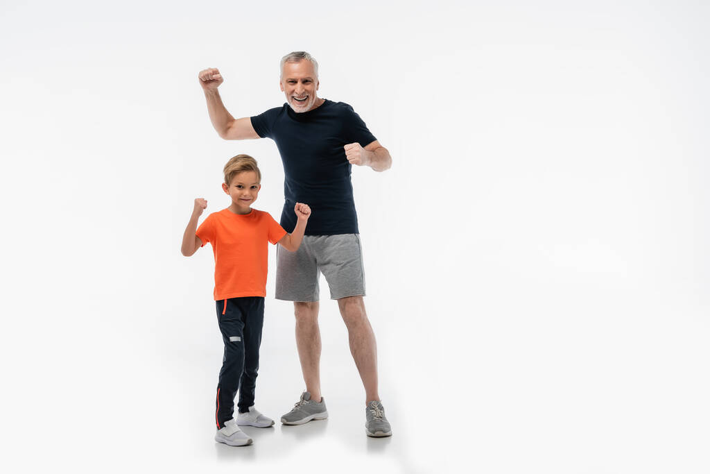 vzrušený dědeček s vnukem ve sportovním oblečení ukazuje vítězství gesto na bílém - Fotografie, Obrázek