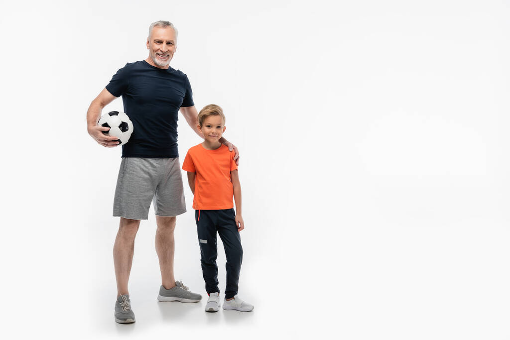 счастливый человек в спортивной одежде, держась за плечо внука, держа футбольный мяч на белом - Фото, изображение
