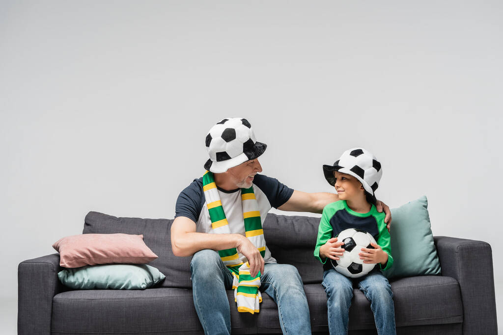 χαρούμενα παππούς και εγγονός κοιτάζοντας ο ένας τον άλλον, ενώ κάθεται στον καναπέ στο ποδόσφαιρο καπέλα ανεμιστήρα απομονώνονται σε γκρι - Φωτογραφία, εικόνα