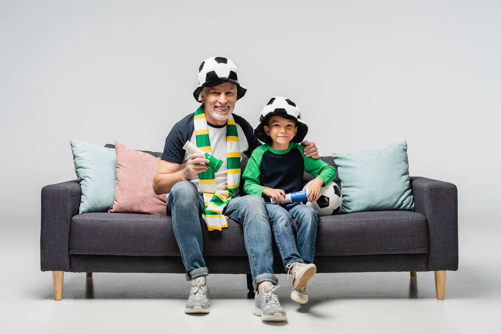 glücklicher Mann und Enkel in Fußballfanhüten mit Hörnern, während sie Fußballspiel auf grau schauen - Foto, Bild
