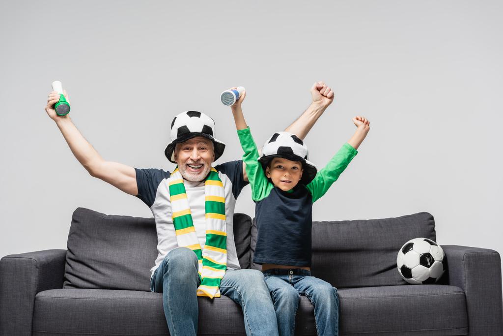 孫を持つ興奮した男は、灰色で隔離されたファンの帽子でサッカー選手権を見ている間に勝つジェスチャーを示す - 写真・画像