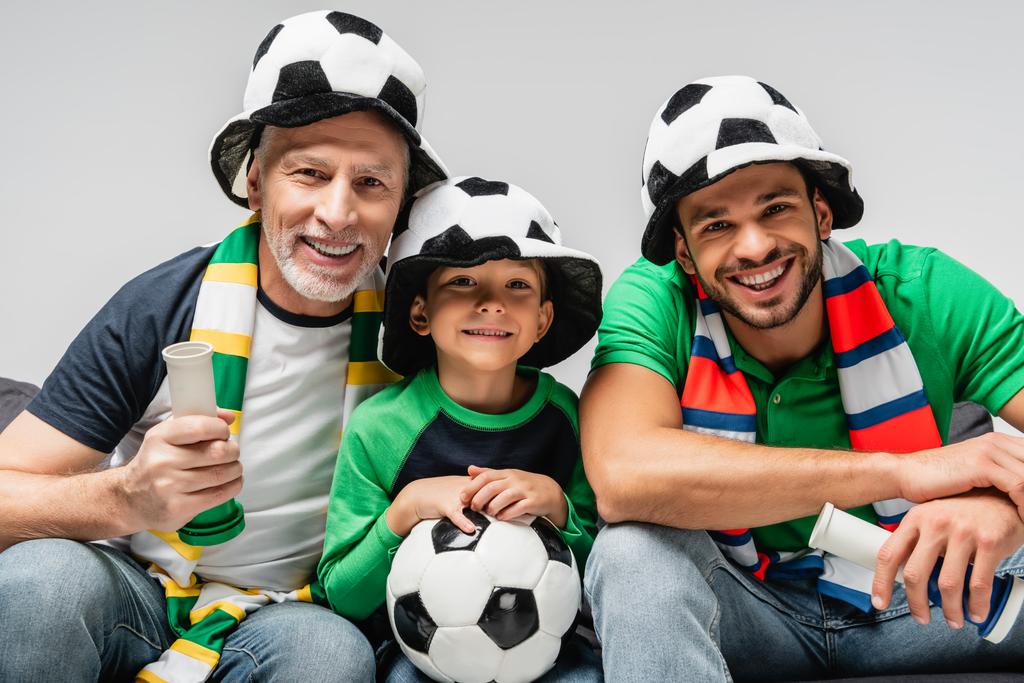 glücklicher Junge mit Fußball, der zusammen mit Papa und Großvater die Fußballmeisterschaft verfolgt, isoliert auf grau - Foto, Bild