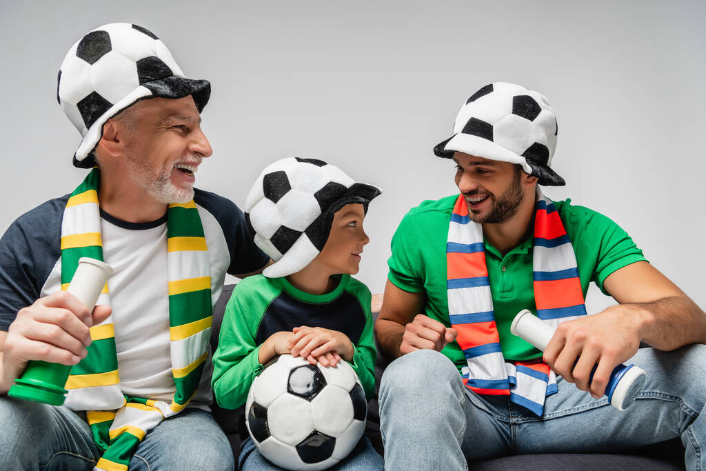 Taraftar şapkalı mutlu adamlar çocuğun yanında boynuzlarını tutuyorlar. Futbol topu gride izole edilmiş. - Fotoğraf, Görsel
