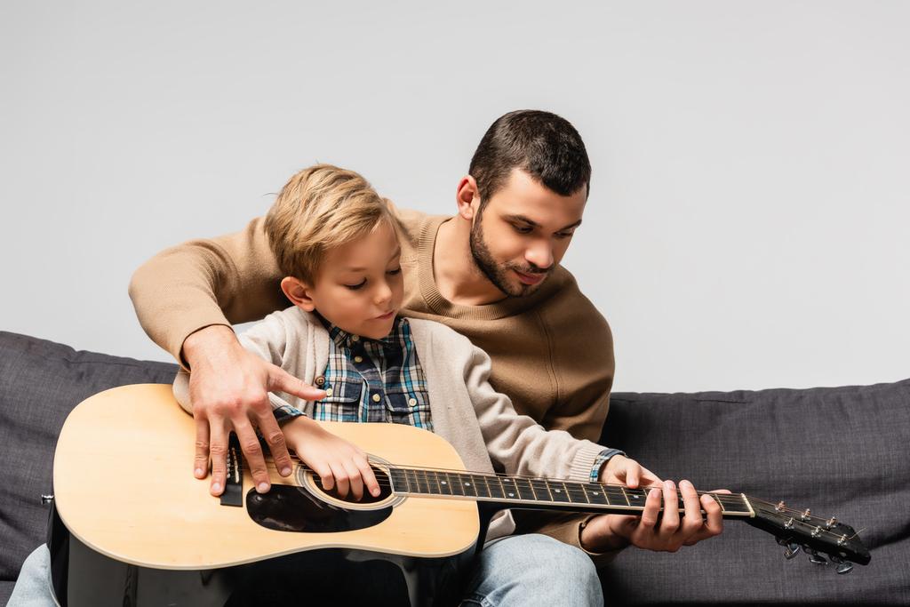 Vater bringt Sohn Akustikgitarre bei, während er isoliert auf grauem Sofa sitzt - Foto, Bild