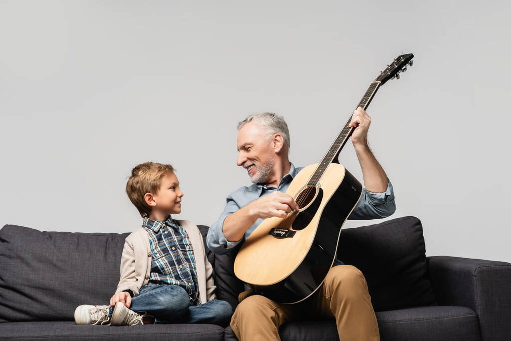 abuelo feliz tocando la guitarra acústica cerca de nieto sonriente aislado en gris - Foto, imagen