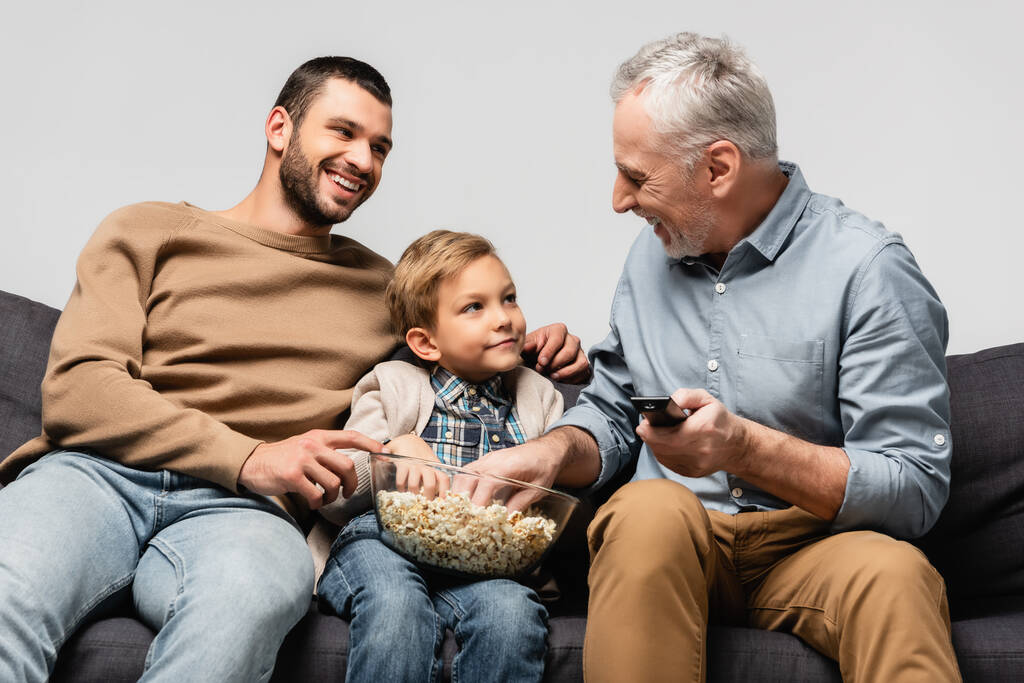 szczęśliwy Dojrzały mężczyzna siedzi na kanapie z pilotem w pobliżu syna i wnuka z miską popcornu odizolowany na szarości - Zdjęcie, obraz