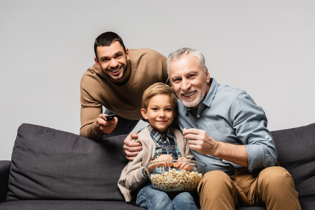 ευτυχισμένος άνθρωπος κρατώντας τηλεχειριστήριο κοντά στον πατέρα και το γιο βλέποντας τηλεόραση και τρώγοντας ποπ κορν απομονωμένο στο γκρι - Φωτογραφία, εικόνα