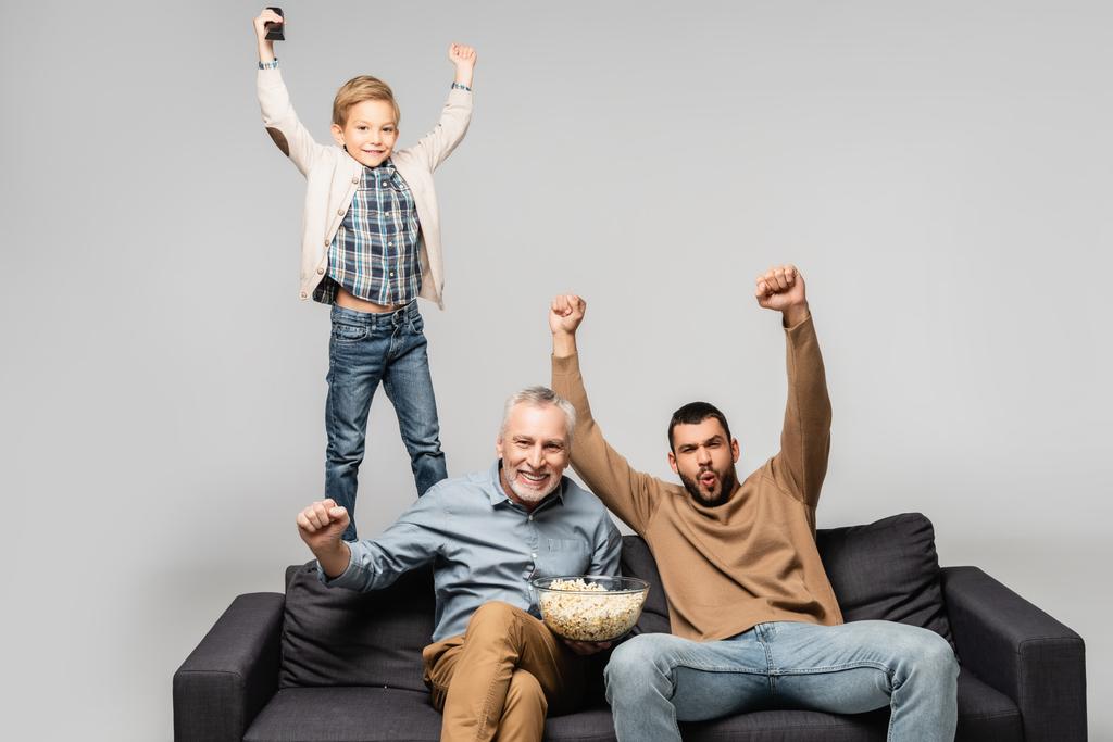 uomini eccitati con ragazzo mostrando vincere gesto mentre si guarda la tv sul divano isolato sul grigio - Foto, immagini