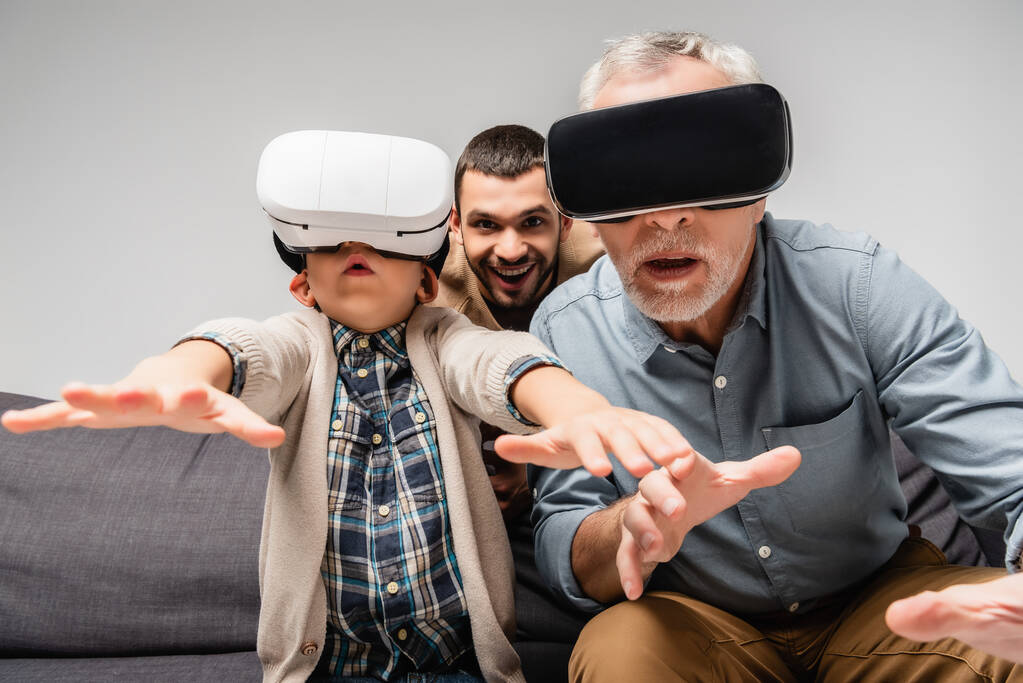 grandfather and grandson gesturing in vr headsets near excited man on background isolated on grey - Photo, Image