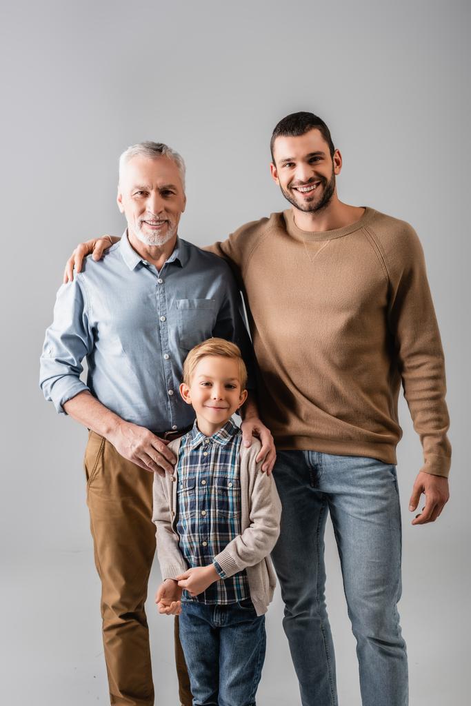 счастливый мальчик, смотрящий в камеру рядом с отцом и дедушкой, изолированные на сером - Фото, изображение