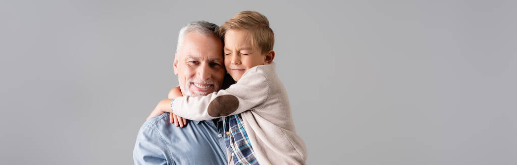 gelukkig volwassen man kijken naar camera terwijl het houden van kleinzoon omarmen hem met gesloten ogen geïsoleerd op grijs, banner - Foto, afbeelding