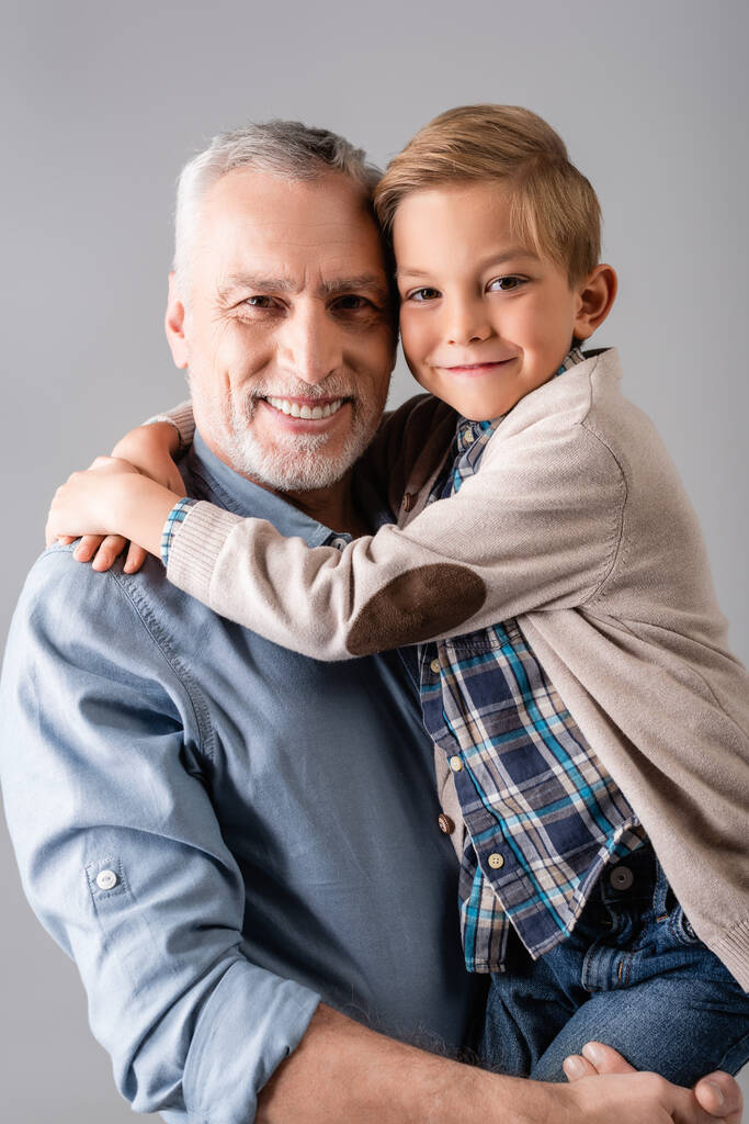 veselý muž usmívá na kameru, zatímco drží šťastný vnuk objímající ho izolované na šedé - Fotografie, Obrázek