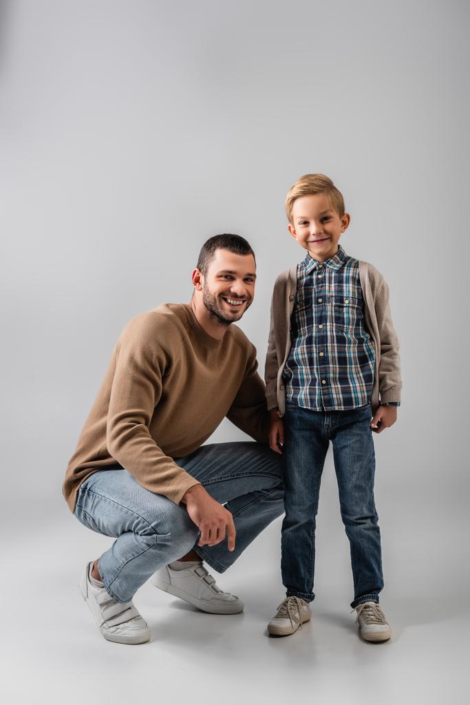 счастливый отец приседает рядом с веселым сыном, улыбаясь вместе в камеру на серой - Фото, изображение