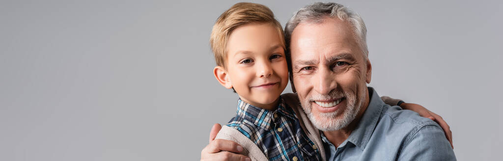 feliz abuelo y nieto abrazándose mientras sonríe a la cámara aislada en gris, pancarta - Foto, Imagen